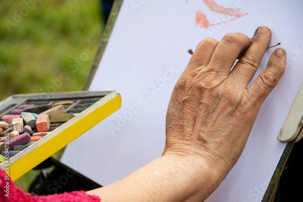 Fototapeta Street artist holding a box with multicolored crayons and pencils for drawing.Close up photography of hands and drawing supplies.He drawing behind the easel with white paper.