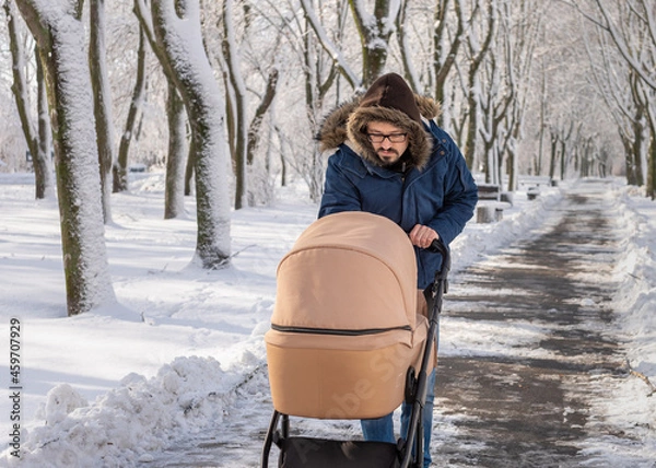 Obraz A bearded dad walking with baby carriage in winter park. A man with a baby stroller walks in a winter snow-covered city park in the cold. Caring father admires baby sleeping in stroller.
