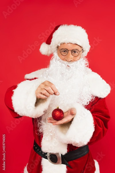 Obraz Santa claus in costume and eyeglasses holding christmas ball isolated on red
