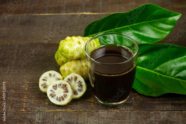 Fototapeta Noni juice in transparent glass and noni fruit with green leaf on wooden background. 