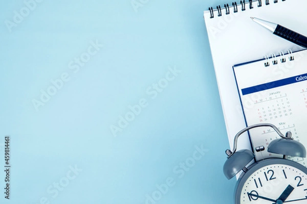 Fototapeta pen with white spiral notepad  and alarm clock calendar placed on blue background , business meeting agenda schedule, travel planning booking or a project event milestone and reminder concept.