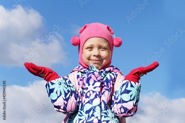Obraz Cute little girl on a background of blue sky with raised hands. Pink clothes. Child outside in winter in cold weather