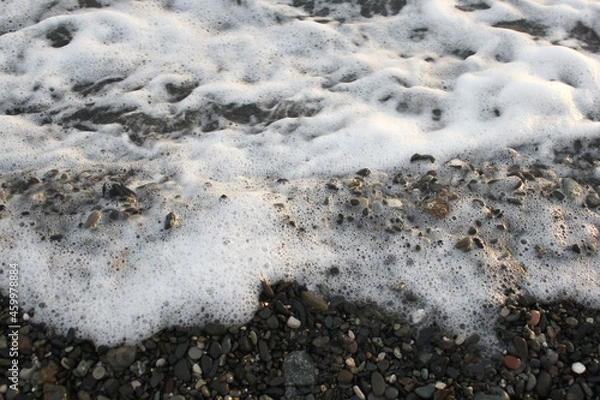 Fototapeta White sea foam on black sand. Sea coast, pebble beach background. Close-up photos, selective focus.