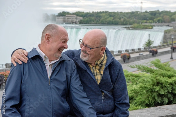 Fototapeta An elderly gay couple sit on a stone wall with Niagara Falls in the background.  They are looking at each other and laughing.  One has his arm around the shoulder of his husband.