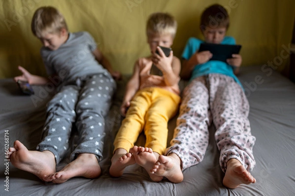 Fototapeta three funny boys lie on couch and play on smartphones and tablets. Children are passionate about game and their devices