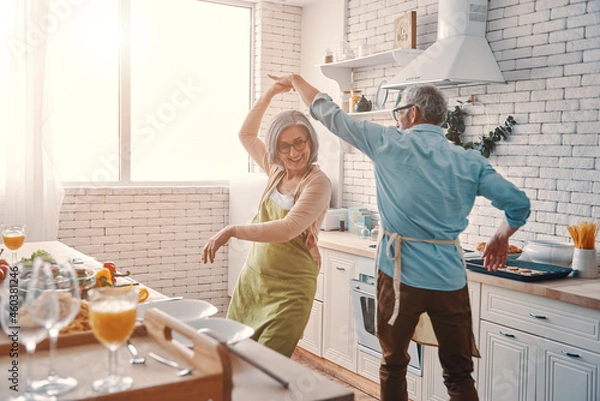 Fototapeta Beautiful playful senior couple in aprons dancing and smiling while preparing healthy dinner at home