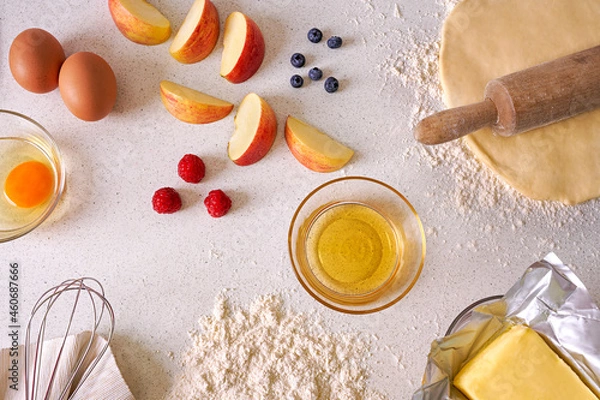 Obraz Table top view displaying raspberries, blueberries, apples, eggs, dough, butter and honey on a light kitchen counter surface