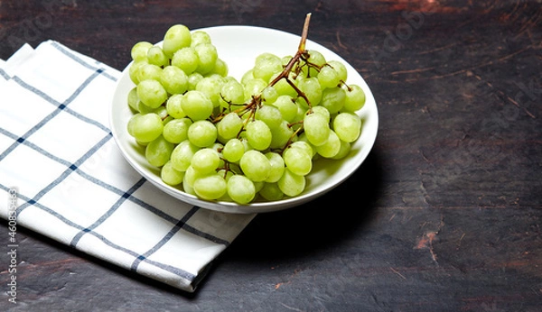Fototapeta Branch of ripe green grape on plate with water drops. Juicy grapes on wooden background, closeup. Grapes on dark kitchen table with copy space