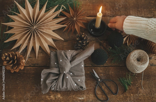 Fototapeta Christmas advent. Hand holding candle and stylish christmas gift wrapped in fabric, scissors, paper star, ornaments on rustic wooden table. Atmospheric moody image, nordic style. Merry Christmas!