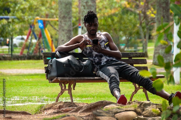Obraz African black man in sunny afternoon, sitting on bench, using smartphone, with his black bag beside him, relaxing outdoors in park, healthy life.