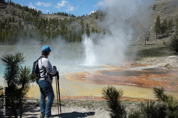 Fototapeta Yellowstone National Park - Hike to Imperial Geyser. Woman standing in front of geyser looking at it erupting. Shot from behind woman. Yellow, gold, orange, and turquoise colors.