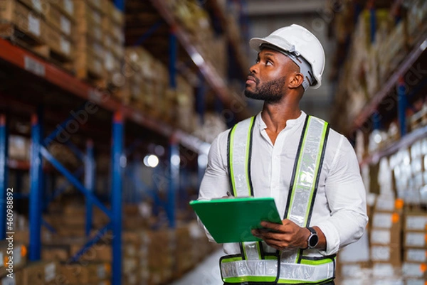 Obraz Portrait of an African warehouse manager holding a clipboard checking inventory in a large distribution center.
