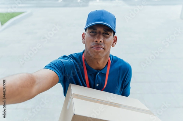Fototapeta Young delivery man looking at camera while calling intercom to deliver package.