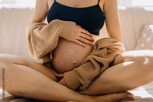 Fototapeta Closeup of a pregnant woman sitting on a sofa in soft light, hugging her belly, wearing a cozy cardigan.