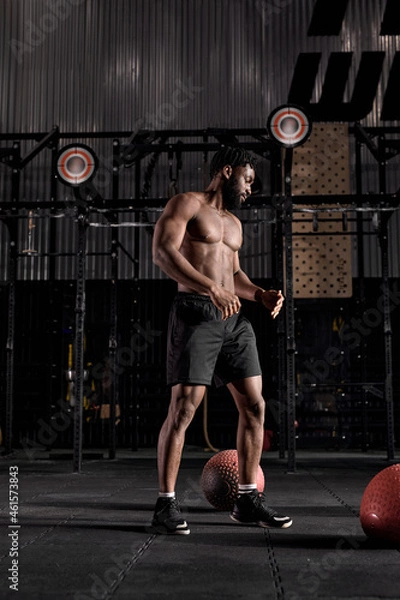 Fototapeta Portrait of handsome muscular male with naked torso posing standing after workout in gym, looking confident and motivated. handsome guy with beard wearing black shorts looking down