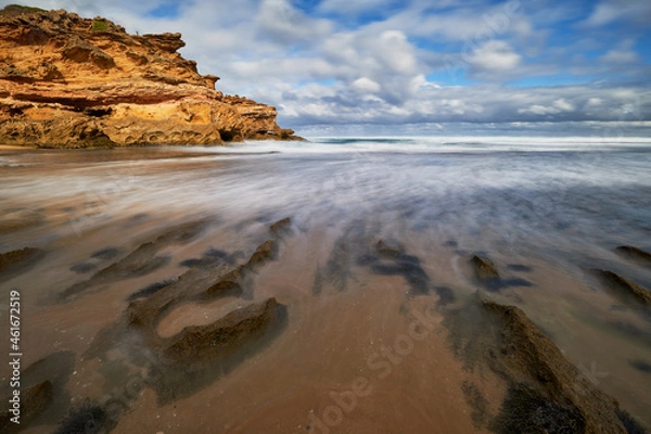 Fototapeta Long exposure of waves washing into a sandy beach shore, with rocky coastline in the background and cloudy blue sky on the horizon, in Victoria, Australia.
