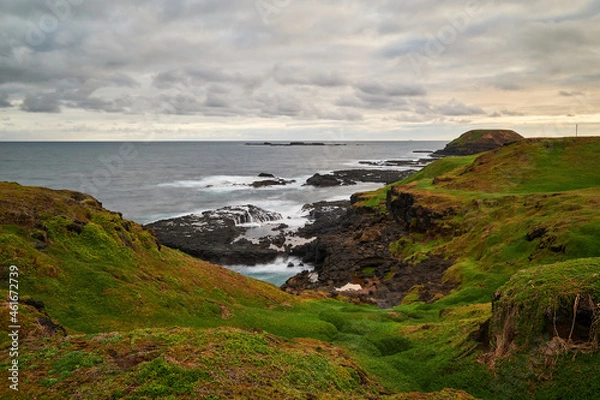 Fototapeta A scenic view overlooking green grassy hills with beautiful volcanic, rocky coastline in the background, deep blue sea and cloudy sky on the horizon, at Phillip Island in Australia.
