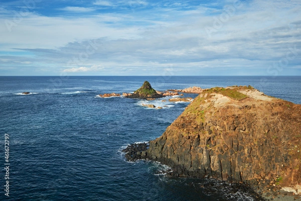 Fototapeta Overlooking a scenic view of a pyramid shaped rocky island with rugged coastline, deep blue sea in the background and cloudy blue sky on the horizon, photographed at Phillip Island, Australia.
