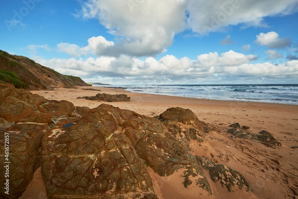 Fototapeta Scenic view of a sandy beach landscape with volcanic rocks in the foreground and ocean in the background, cloudy blue sky above the horizon at Phillip Island, Australia.
