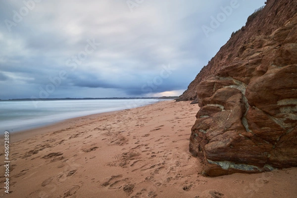 Fototapeta Scenic view of a sandy beach landscape with volcanic rocks in the foreground and ocean in the background, cloudy blue sky above the horizon at Phillip Island, Australia.
