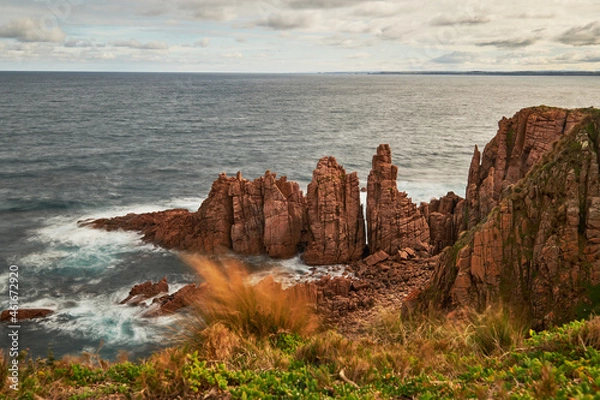 Fototapeta A scenic view overlooking waves breaking against red pillars of rock with the vast ocean on the horizon, in the background is the coastline and sea with cloudy sky above, at Phillip Island Australia.
