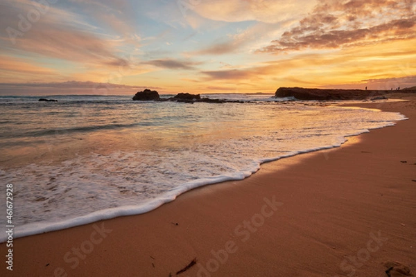Fototapeta A beautiful beach sunset illuminating the sky with vibrant orange colours, a scenic view of the ocean with waves washing onto the shore along the coast of Phillip Island, Australia.

