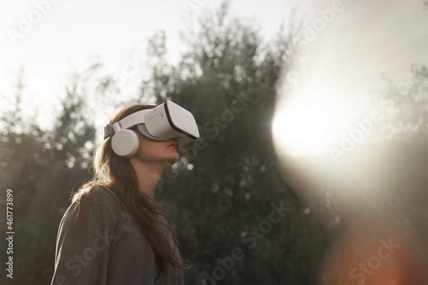 Fototapeta The girl, wearing virtual reality glasses, holds the glasses with her hands. Against the background of green trees. There is a beam of sunlight nearby. High quality photo