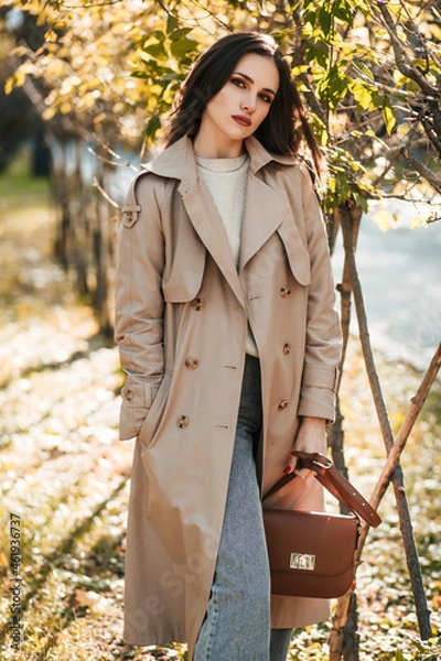 Fototapeta A beautiful tall girl of model appearance in a beige raincoat and gray jeans walks in a city park on a warm autumn day.Lifestyle concept. Autumn street fashion look.
