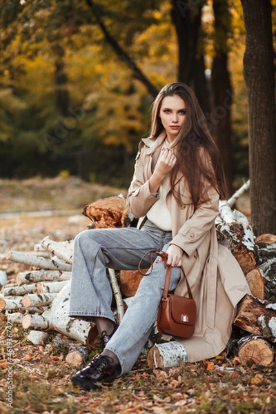 Fototapeta A beautiful tall girl of model appearance in a beige raincoat and gray jeans sits on sawn logs in a city park on a warm autumn day. Autumn street trendy look.