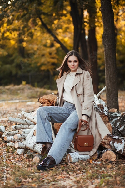 Fototapeta A beautiful tall girl of model appearance in a beige raincoat and gray jeans sits on sawn logs in a city park on a warm autumn day. Autumn street trendy look.