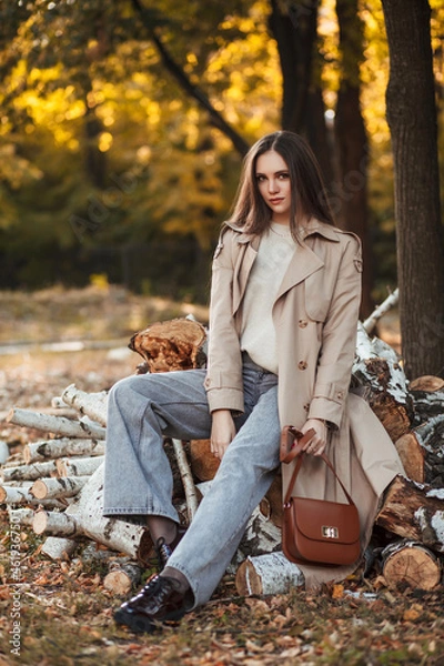 Fototapeta A beautiful tall girl of model appearance in a beige raincoat and gray jeans sits on sawn logs in a city park on a warm autumn day. Autumn street trendy look.