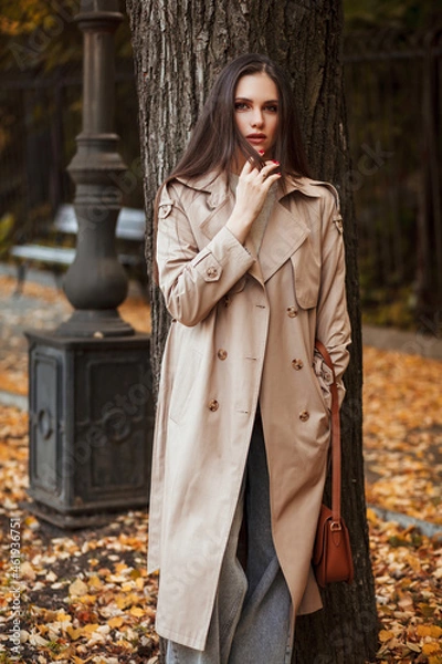 Fototapeta A beautiful tall girl of model appearance in a beige raincoat and gray jeans walks in a city park on a warm autumn day.Lifestyle concept. Autumn street fashion look.