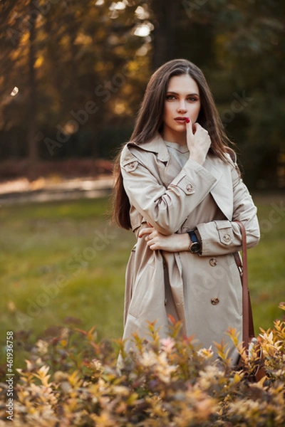 Fototapeta A beautiful tall girl of model appearance in a beige raincoat and gray jeans walks in a city park on a warm autumn day.Lifestyle concept. Autumn street fashion look.