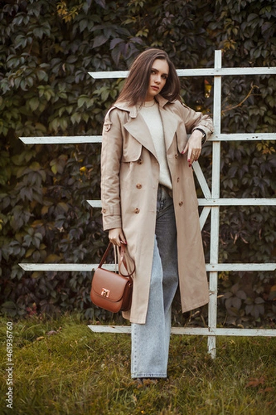 Fototapeta A beautiful tall girl of model appearance in a beige raincoat and gray jeans walks in a city park on a warm autumn day.Lifestyle concept. Autumn street fashion look.