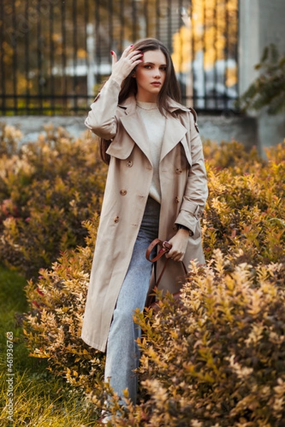 Fototapeta A beautiful tall girl of model appearance in a beige raincoat and gray jeans walks in a city park on a warm autumn day.Lifestyle concept. Autumn street fashion look.