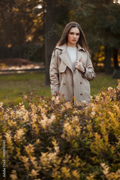 Fototapeta A beautiful tall girl of model appearance in a beige raincoat and gray jeans walks in a city park on a warm autumn day.Lifestyle concept. Autumn street fashion look.