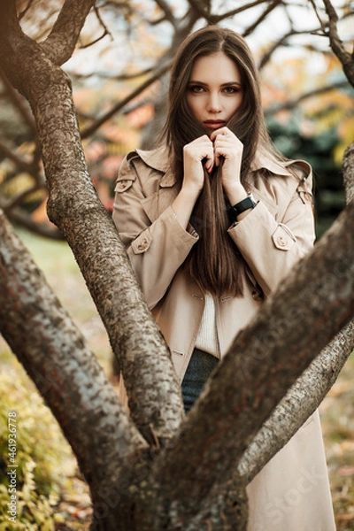 Fototapeta A beautiful tall girl of model appearance in a beige raincoat and gray jeans walks in a city park on a warm autumn day.Lifestyle concept. Autumn street fashion look.