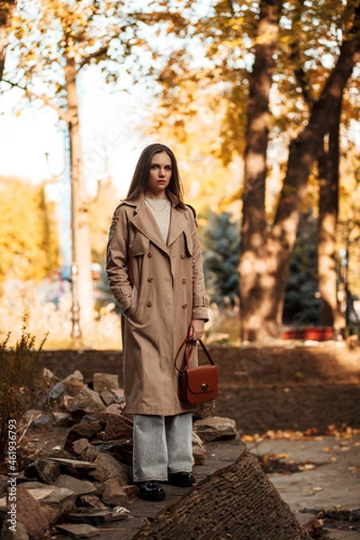 Fototapeta A beautiful tall girl of model appearance in a beige raincoat and gray jeans walks in a city park on a warm autumn day.Lifestyle concept. Autumn street fashion look.