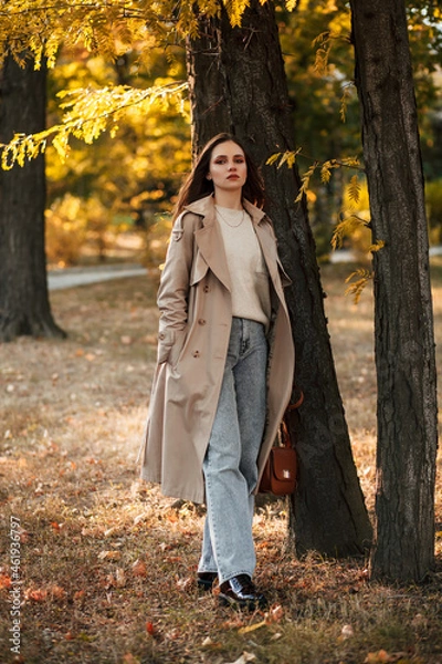 Fototapeta A beautiful tall girl of model appearance in a beige raincoat and gray jeans walks in a city park on a warm autumn day.Lifestyle concept. Autumn street fashion look.