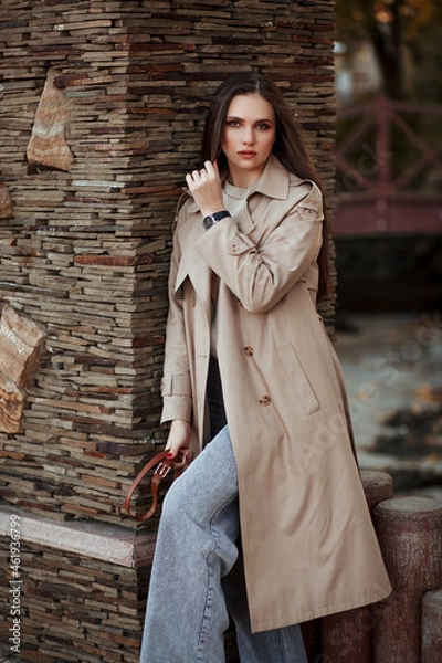 Fototapeta A beautiful tall girl of model appearance in a beige raincoat and gray jeans walks in a city park on a warm autumn day.Lifestyle concept. Autumn street fashion look.