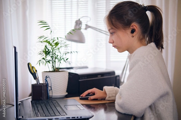Fototapeta Young beautiful girl works at a laptop, holding a hand on a computer mouse at home at a work table in a bright room.
