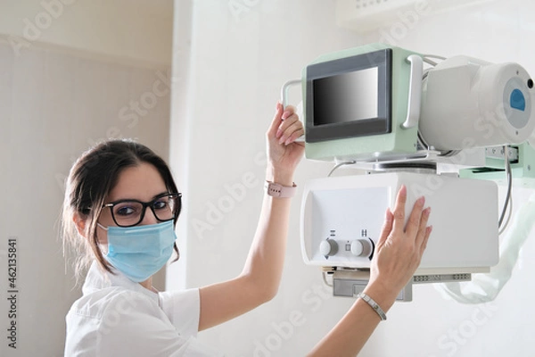 Fototapeta asian woman technician adjusting x-ray machine in a clinic radiology department. young doctor wearing eyeglasses and face mask turning on scanner to make an image.