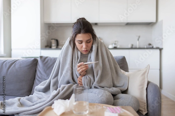 Fototapeta Unhealthy upset young female with fever looking at thermometer with high degree temperature. Sick ill girl suffer from flu or cold, have covid-19 symptoms sitting covered blanket on couch at home.
