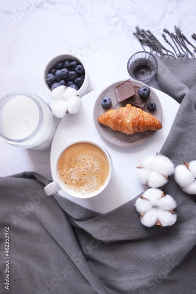 Fototapeta Cup of coffee with croissant, chocolate bar, blueberries, cotton flowers and grey scarf on the white marble background. Autumn, winter coffee break. Cozy warm home latte. Empty place for text. 