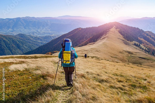 Obraz A girl in colored clothes with a large backpack walks along a beautiful mountain range in the Carpathians during a sunset