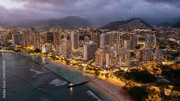 Fototapeta Aerial view of waikiki beach at night in Honolulu, Hawaii. Skyscrapers with lights can be seen with mountains and a sandy beach.
