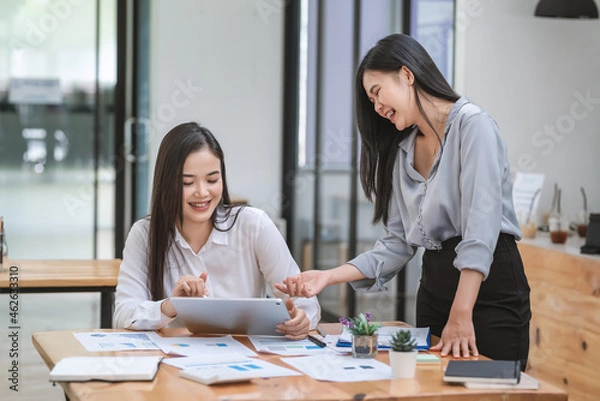 Fototapeta Two beautiful young Asian businesswoman smiling happily working together using a document tablet graph placed at the office.