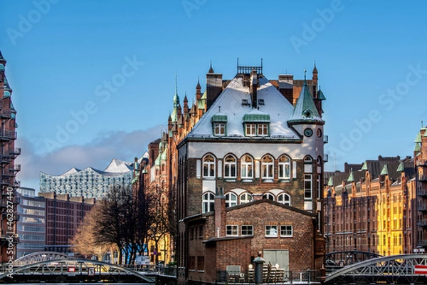 Fototapeta A view on the Speicherstadt with the Wasserschloss and the Elbphilharmonie in Hamburg, on a cold winter day with snow on the roofs and a blue sky