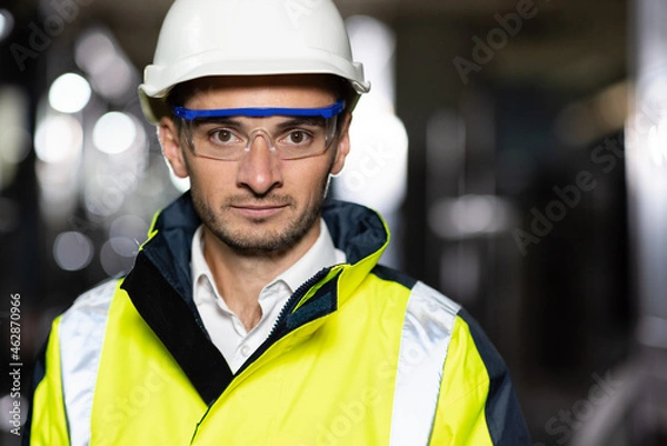 Fototapeta Portrait of Professional Heavy Industry Engineer or Worker Wearing Safety Uniform, Goggles and Hard Hat Looks at Camera. In the Background Unfocused Large Industrial Factory