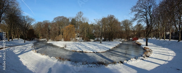 Fototapeta Park of the city of Moers in Germany with an icy brook and snow-covered paths with idyllic blue skies in winter 